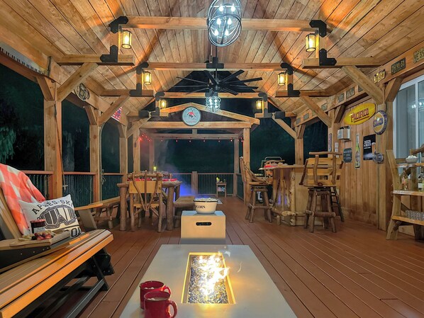 A rustic wooden deck with tables and chairs, a fire feature, and a bar area with signs.