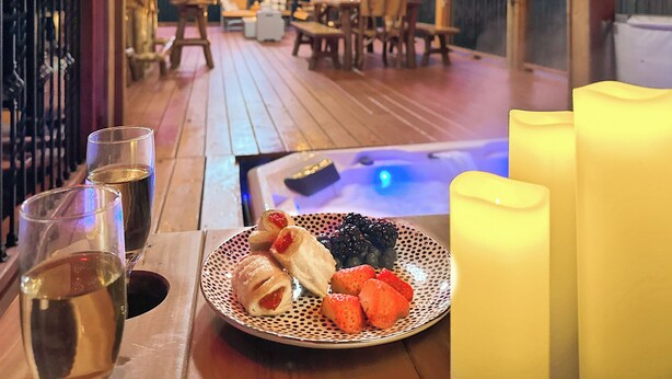 A wooden deck with a hot tub, a dining table with chairs, and a plate of pastries and berries.