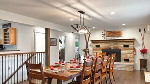 A dining area with a long wooden table set for six, flanked by wooden chairs. A stone fireplace with a modern design is visible in the background.