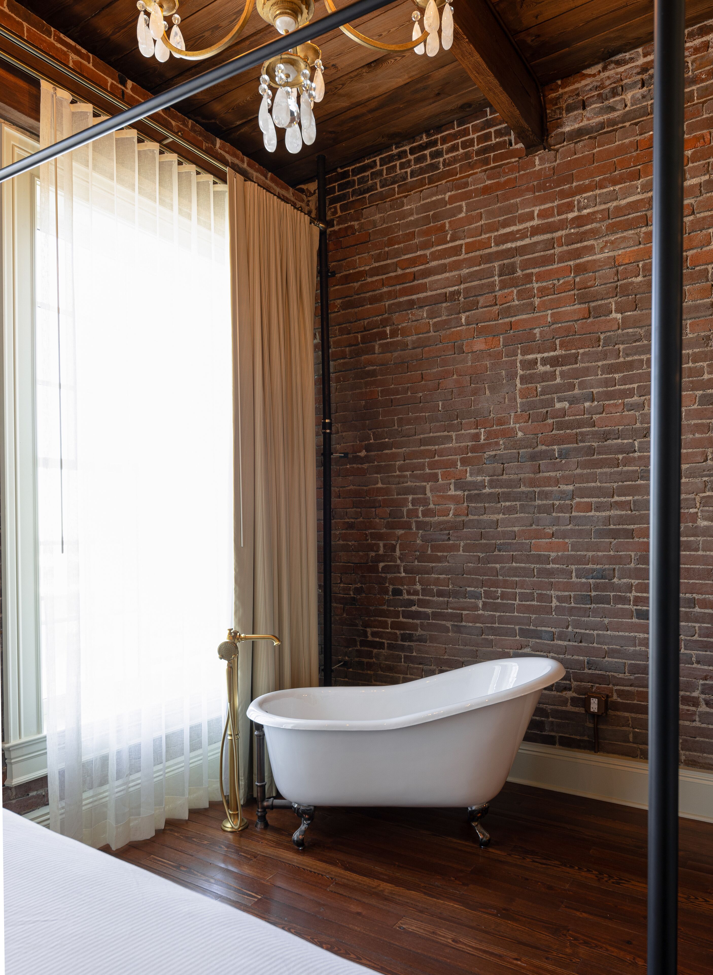 A bathroom with a white freestanding bathtub, a wooden floor, a brick wall, and a chandelier.