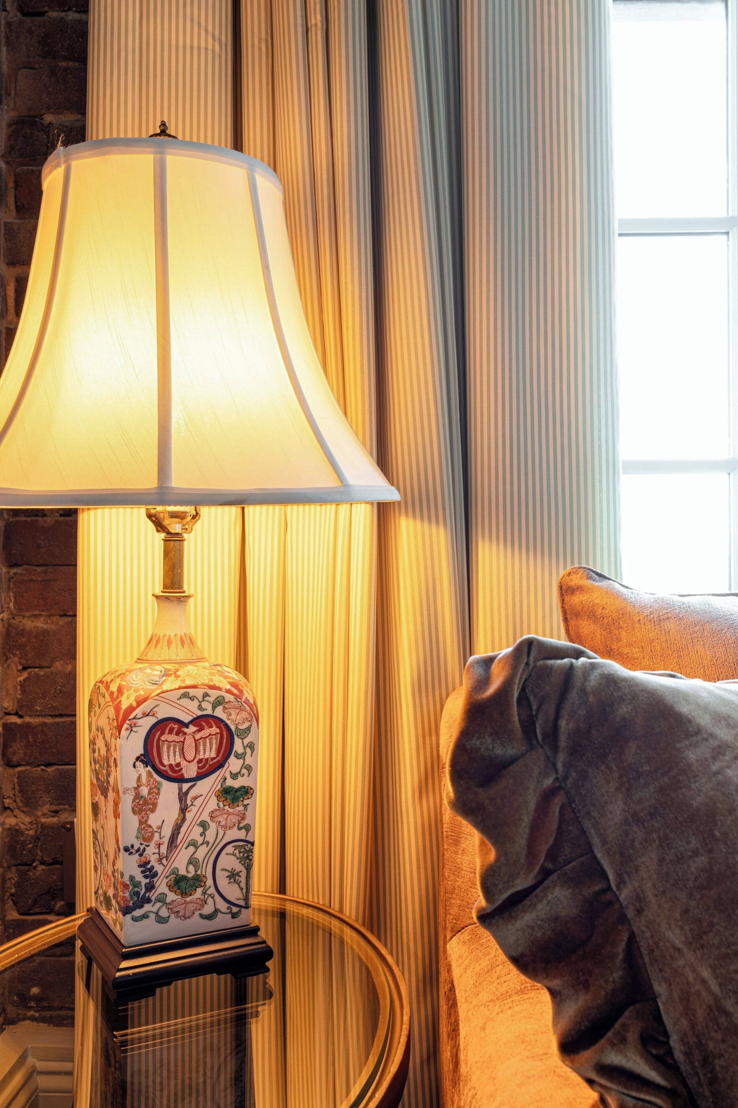 Una lámpara con una base decorativa, un sillón acolchado y una ventana con cortinas.