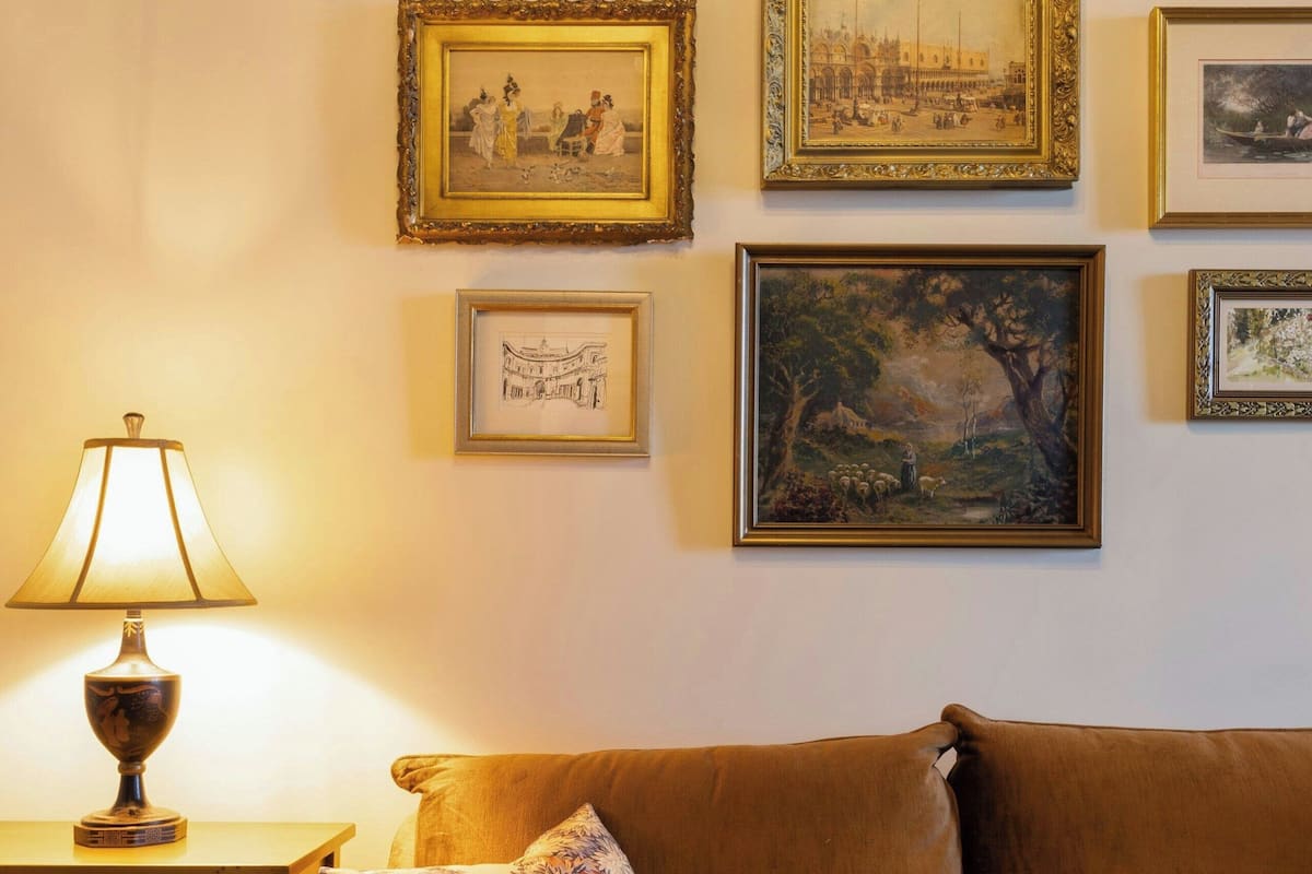 Una sala de estar con un sofá marrón, una mesa con una lámpara y varias obras de arte enmarcadas en la pared.