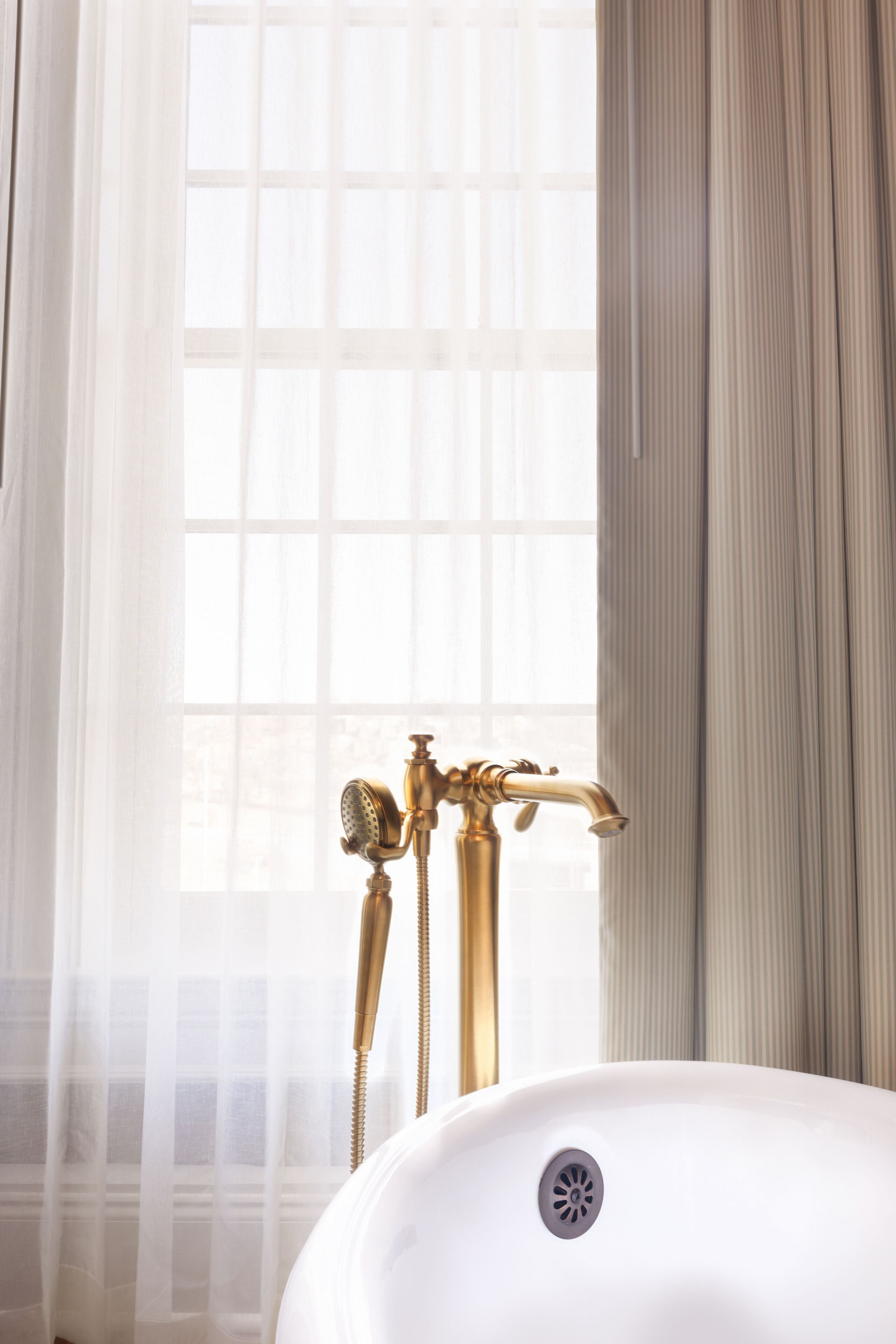 Une salle de bain avec une baignoire blanche, une pomme de douche dorée et une fenêtre ornée de rideaux légers.