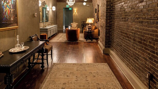 A hallway with a wooden floor, a brick wall, and a chandelier. There is a dining table with chairs and a rug.