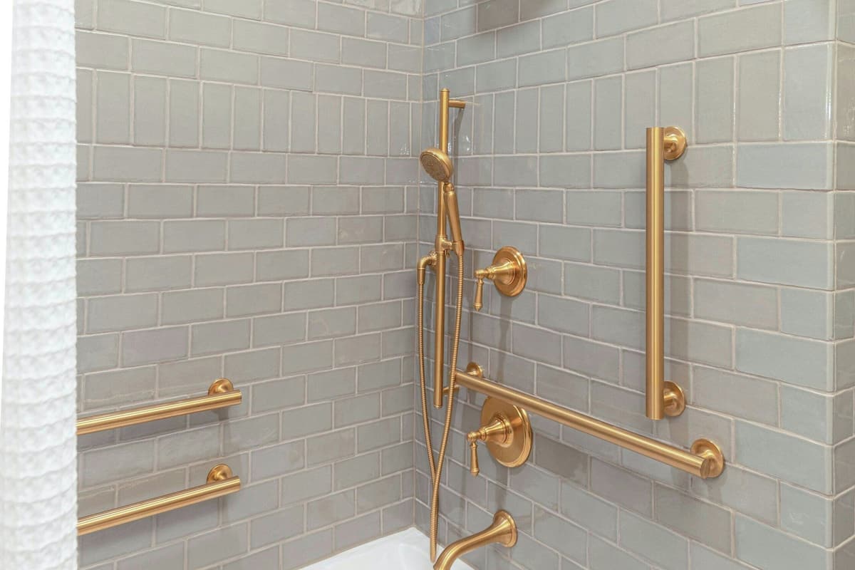 Un baño con una bañera blanca, accesorios dorados y paredes cubiertas de baldosas grises claras.