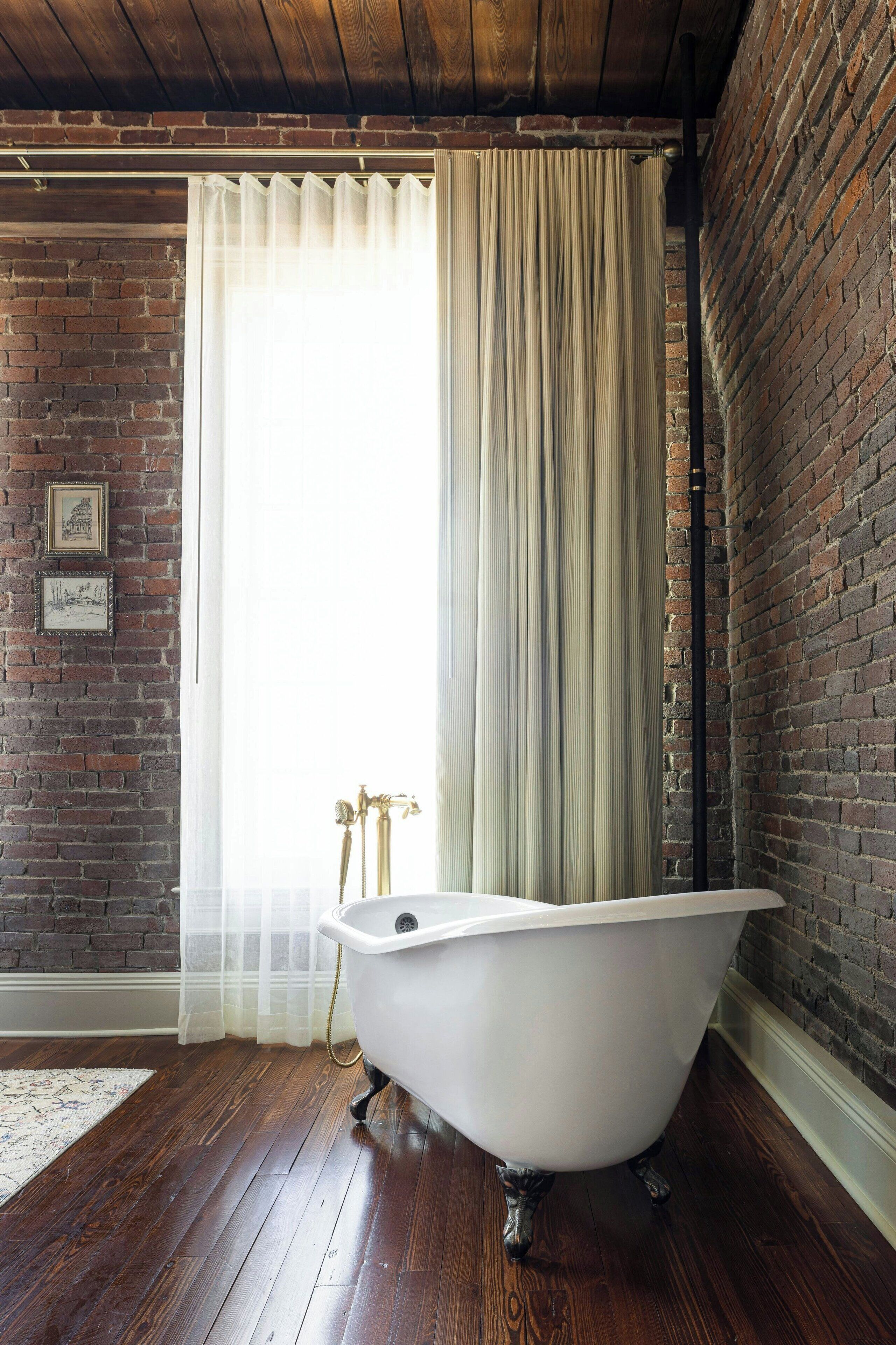 Une pièce avec un mur en briques, une baignoire blanche, un plancher en bois et une fenêtre ornée de rideaux légers.