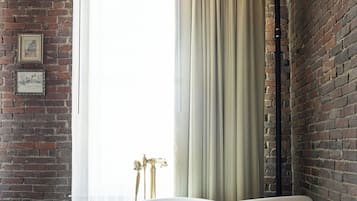 Une pièce avec un mur en briques, une baignoire blanche, un plancher en bois et une fenêtre ornée de rideaux légers.