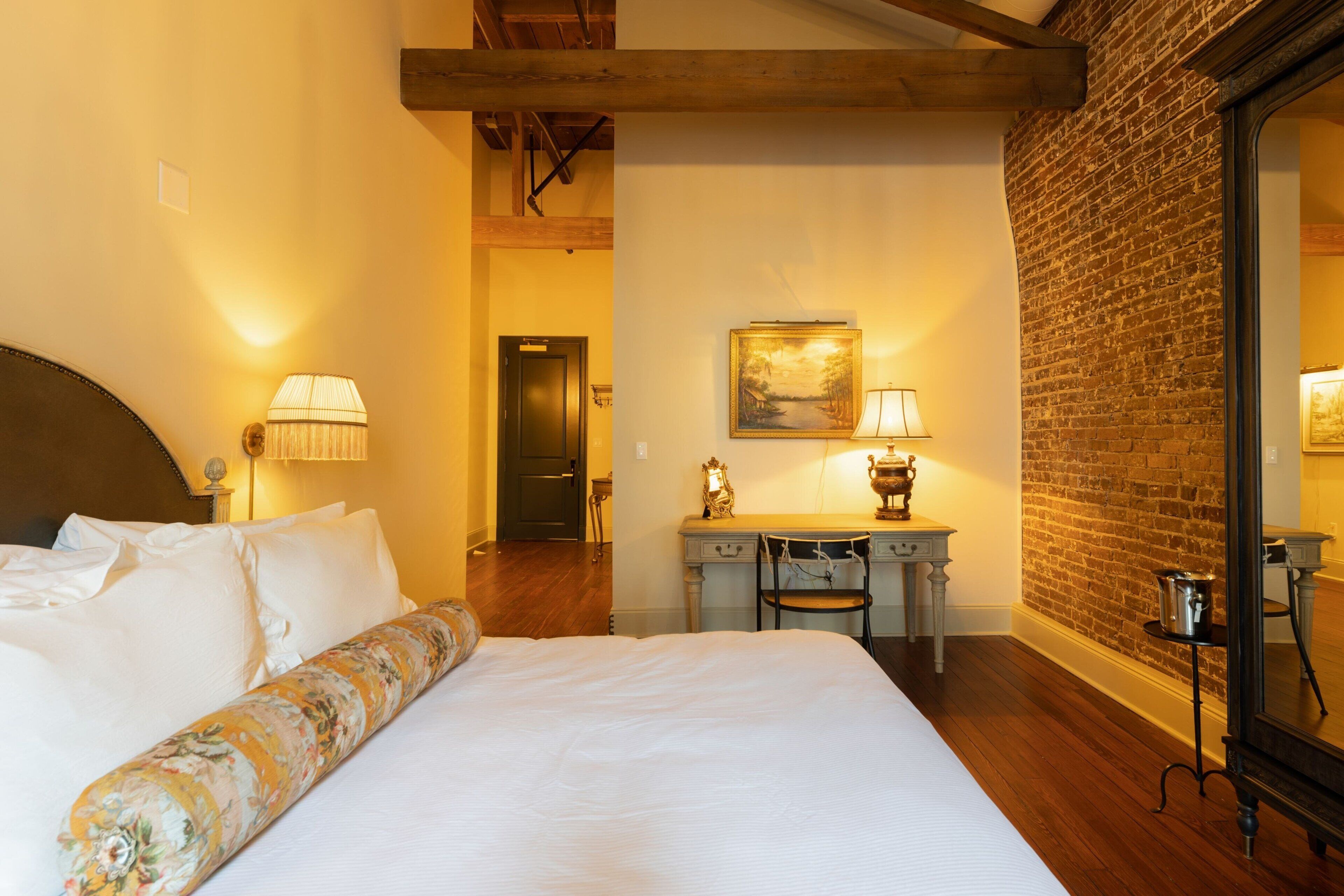 Een hotelkamer met een groot bed, twee bedlampen, een bureau met stoel, een schilderij aan de muur en een bakstenen muur.