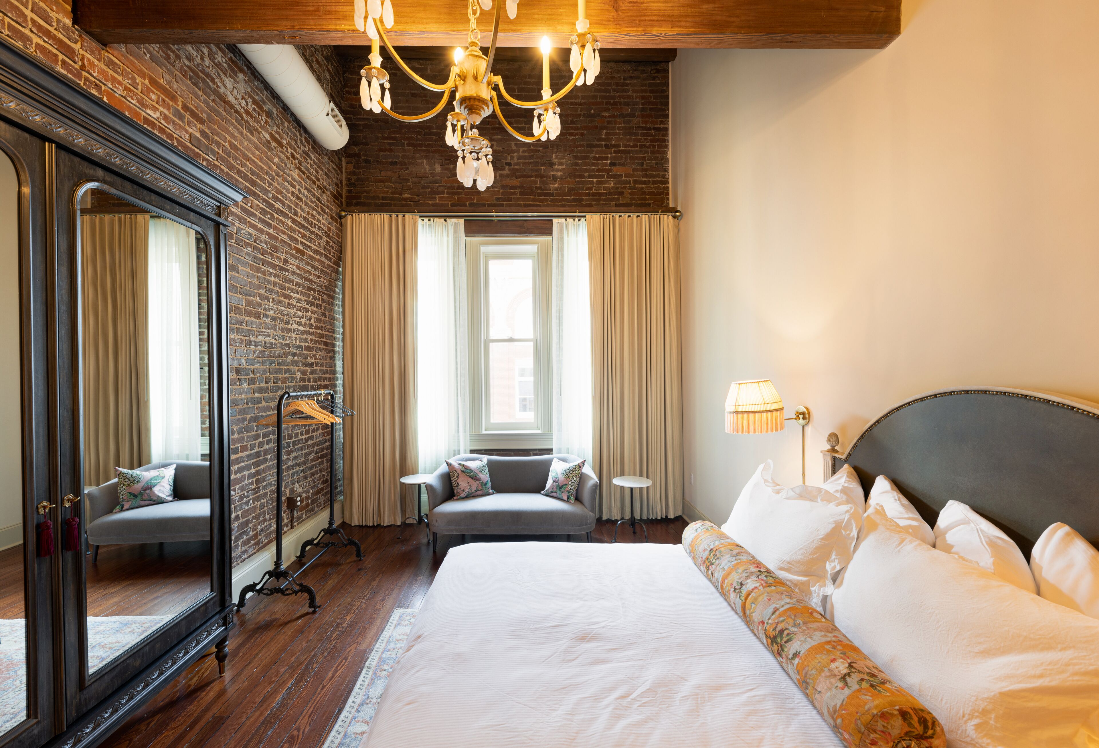 Habitación de hotel con una cama grande, una zona de estar con sofá, un espejo, una araña y vistas de la ciudad a través de la ventana.