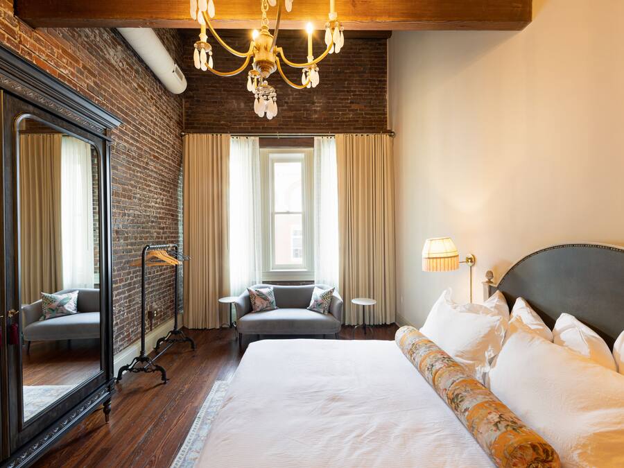 Habitación de hotel con una cama grande, una zona de estar con sofá, un espejo, una araña y vistas de la ciudad a través de la ventana.