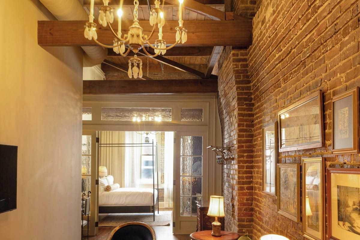 Una habitación de hotel acogedora con techo de madera, una araña, un sofá verde, una pequeña mesa redonda con una lámpara y una cama con vista al exterior.