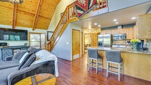 A spacious living area with a high ceiling, wooden beams, a staircase, a kitchen with modern appliances, and a cozy seating area.