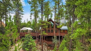 A cabin surrounded by tall pine trees, with a wooden deck and a covered outdoor area.