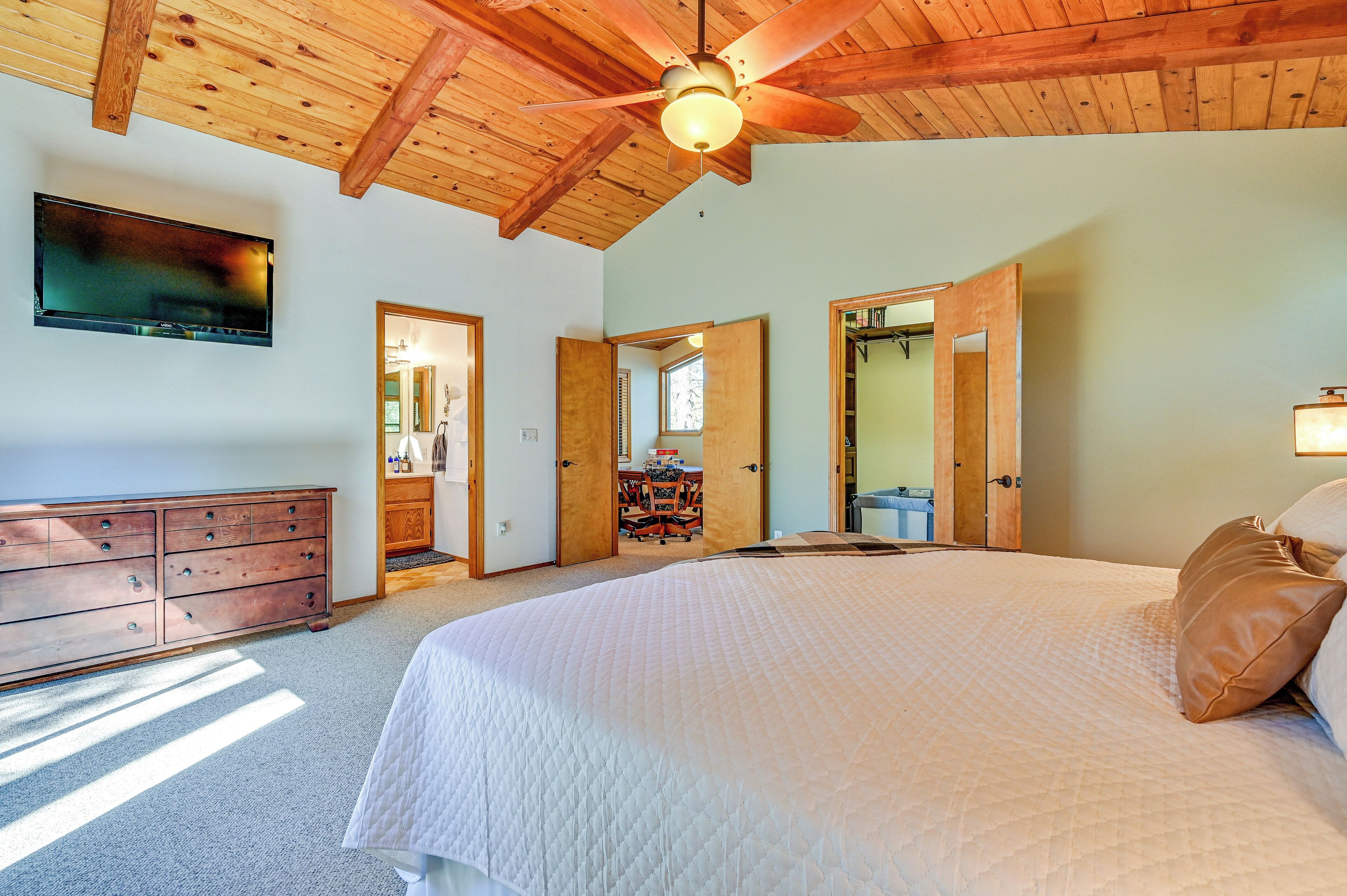 A bedroom with a large bed, wooden ceiling beams, a ceiling fan, a dresser, and a television on the wall.