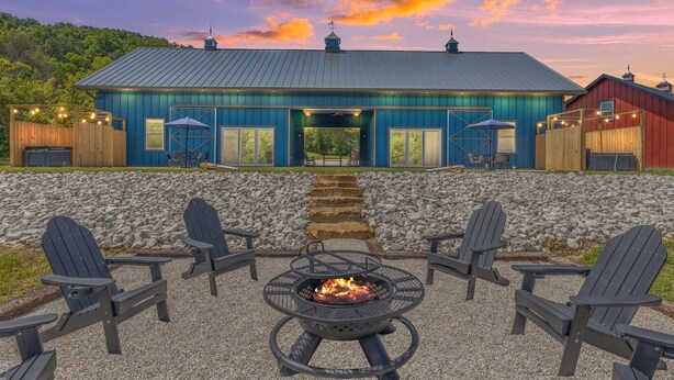 A rustic building with a blue exterior, a fire pit in the center, and Adirondack chairs around it.