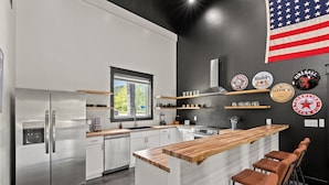 A modern kitchen with a stainless steel refrigerator, white cabinets, a wooden bar counter, and a black accent wall with beer coasters.