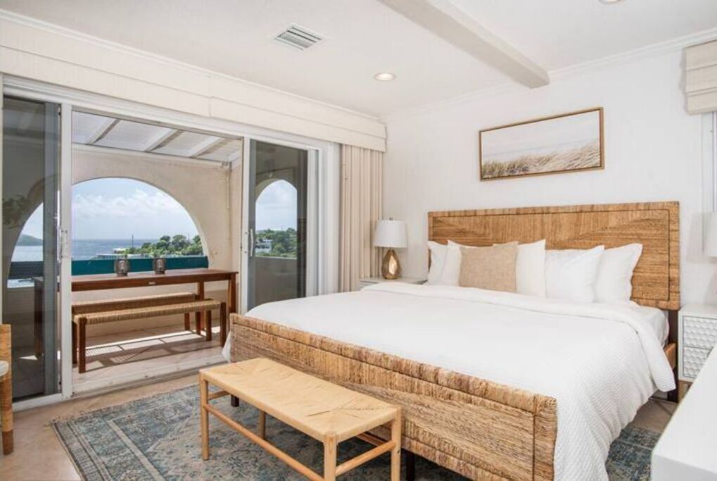A bedroom with a large bed, a wooden headboard, a bench, a table, and a view of the sea through a glass door.