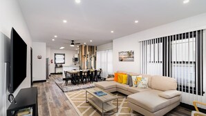 A modern living room with a sofa, coffee table, and TV. There is a dining area with a table and chairs. The room has wooden flooring and a large window with blinds. - Sand & Sun Getaway | Drive to Beach Game Room + 2 Living Areas |Game Room | Minutes to Beach (Michigan City)