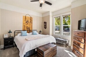 A bedroom with a bed, a wooden bench, a dresser, a television, a window, and a ceiling fan.