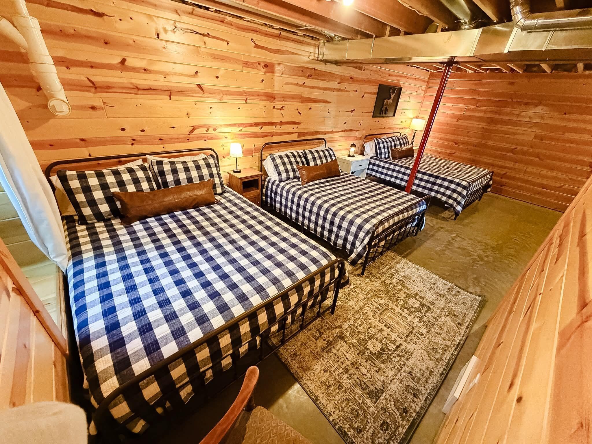 A cozy cabin-style room with three beds, each with checkered bedding, a wooden floor, and a rug.