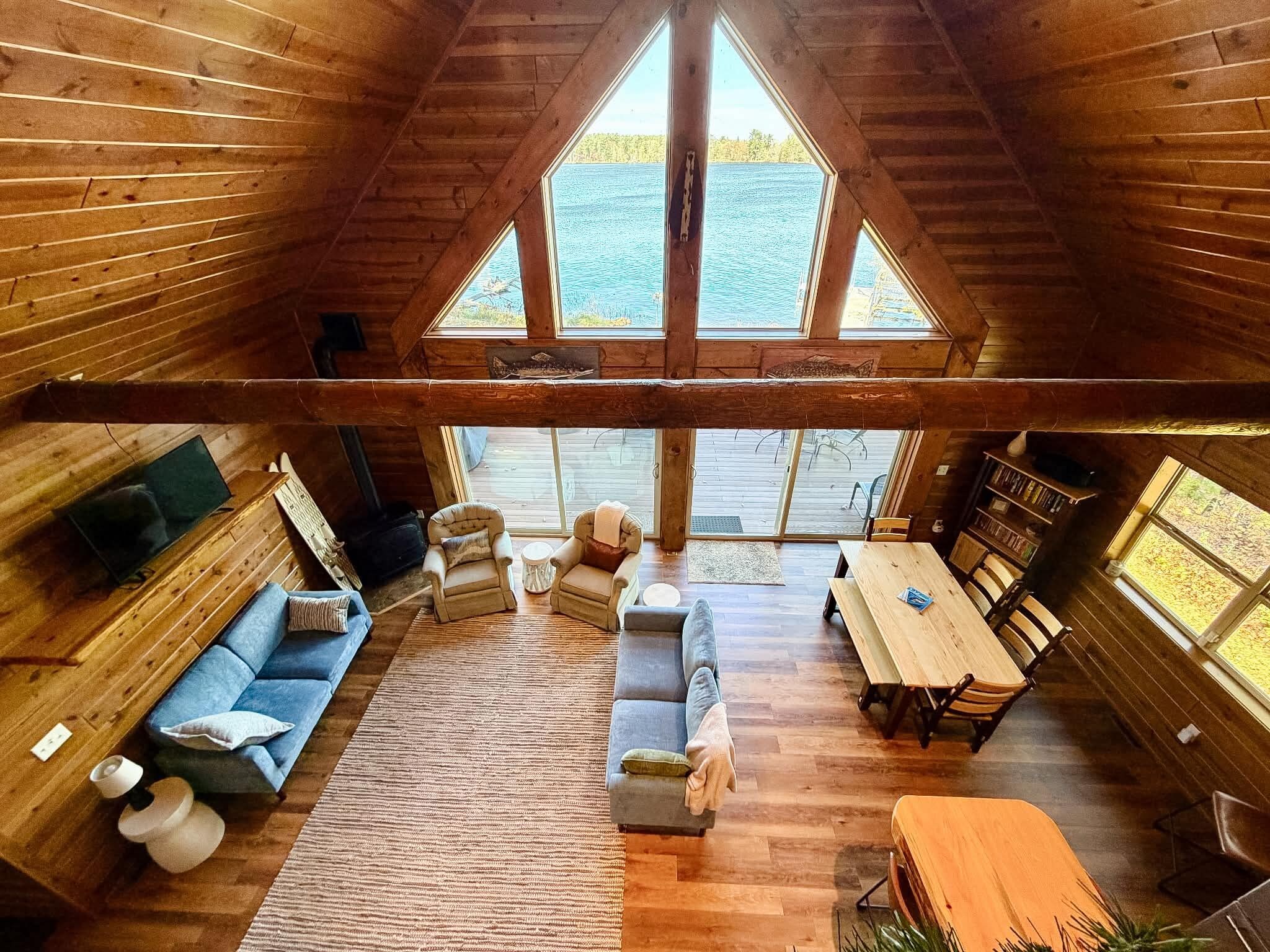 A cabin interior with a loft-style ceiling, wooden walls, a sofa, a dining table, and a view of a lake through large windows.