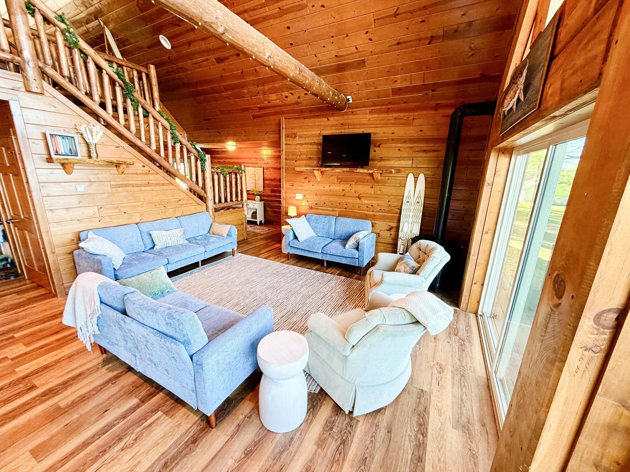 A log cabin interior with a wooden staircase, two sofas, a round coffee table, and a television mounted on the wall.