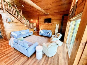 Interior de uma cabana de madeira com escada de madeira, dois sofás, uma mesa de centro redonda e uma televisão montada na parede.