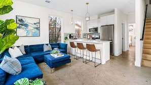 Uma sala de estar moderna com um sofá azul, uma mesa de centro e uma área de cozinha com armários brancos e eletrodomésticos de aço inoxidável.