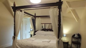 Ein Himmelbett mit weißer Bettwäsche und Kissen, ein Nachttisch mit Lampe sowie ein kleines Regal mit einer Karaffe.