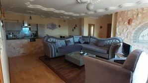 Een woonkamer met een hoekbank, een salontafel en een televisie. Achtergrond is een keuken.