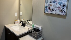 Une salle de bain avec un lavabo blanc, un robinet noir et un miroir. On y trouve des articles de toilette sur le comptoir et une œuvre d’art florale encadrée au mur.