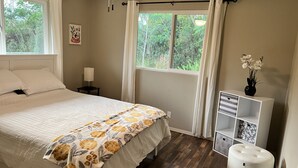 Une chambre à coucher comprenant un lit, une table de chevet, une lampe, une fenêtre avec des rideaux, un ventilateur de plafond et une petite armoire blanche sur laquelle est posé un vase de fleurs.