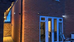 A brick building with a wooden gate, a sliding glass door, and a small patio area with outdoor furniture. - Comfortable 5-Guest Abode -  in Quiet Village (Grantham)