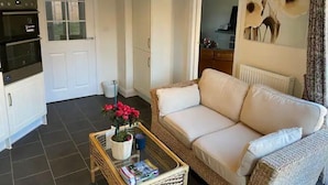 A living room with a sofa, a coffee table with magazines and a plant, a kitchen area with appliances, and a door leading to another room. - Comfortable 5-Guest Abode -  in Quiet Village (Grantham)