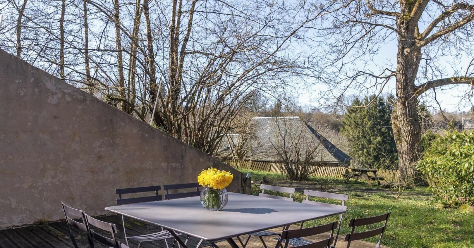 Un patio con una mesa y sillas, un florero con flores amarillas y vistas a árboles y una valla de madera.