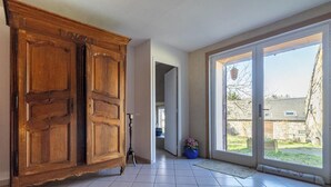 A room with a large wooden wardrobe, a door leading to a garden, and a view of a stone building outside.