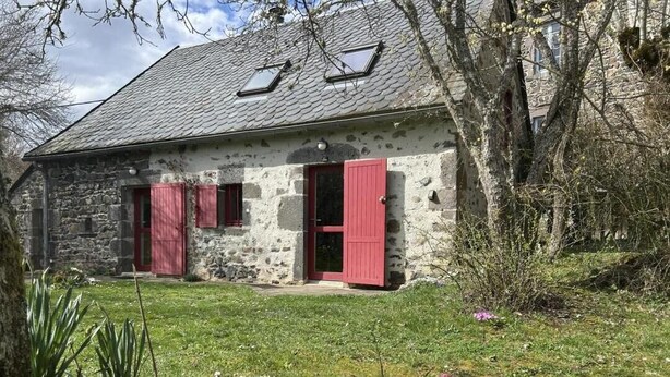 A stone building with red shutters, a grassy yard, and flowering plants.
