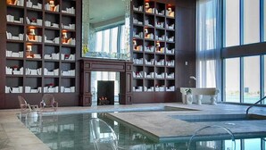 Uma ampla área de piscina interna com paredes de vidro transparente, ladeada por estantes de livros e uma lareira.