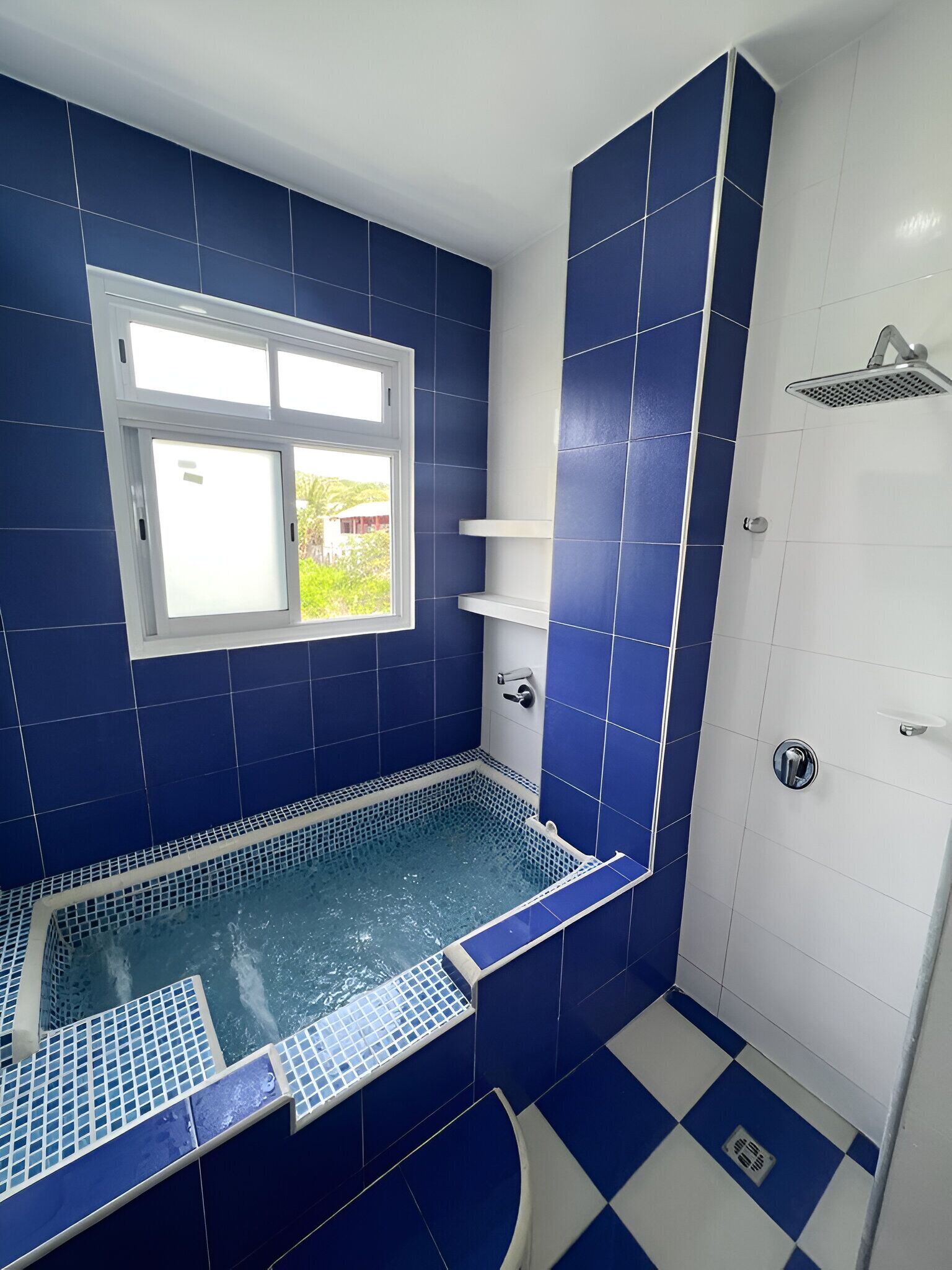 A bathroom with a blue-tiled shower, a white bathtub, and a window with a view of greenery.