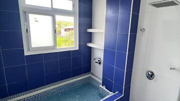 A bathroom with a blue-tiled shower, a white bathtub, and a window with a view of greenery.