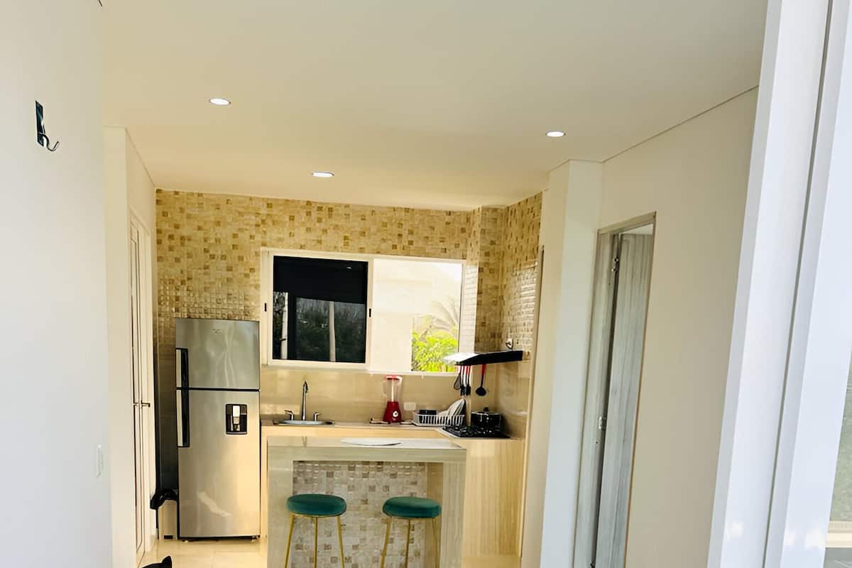 A compact living space with a kitchenette, a black sofa, two bar stools, and a small white table.