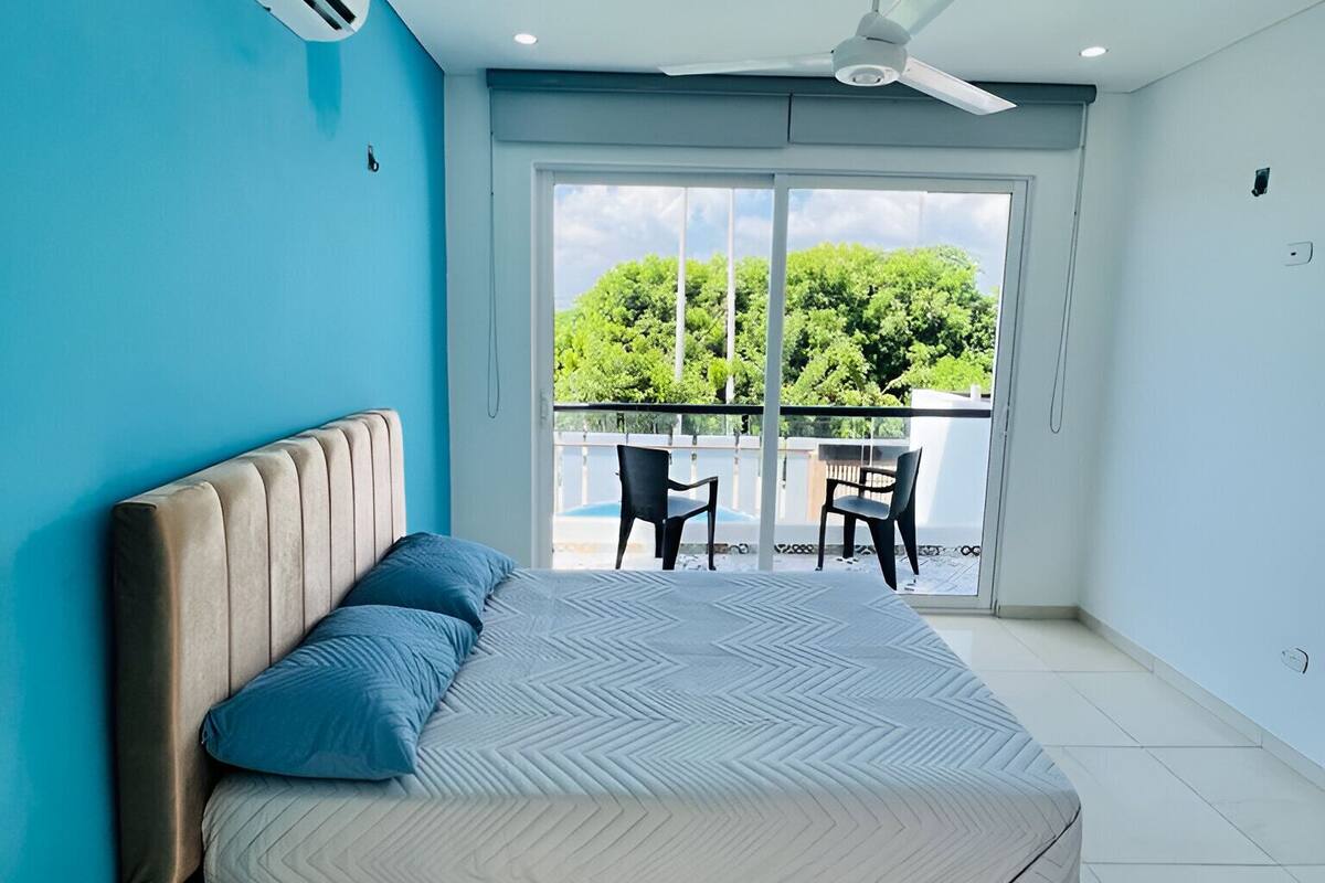A bedroom with a large bed, an air conditioning unit, a ceiling fan, and a view of a balcony with chairs.