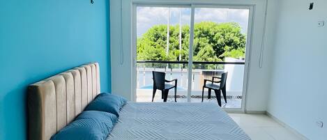 A bedroom with a large bed, an air conditioning unit, a ceiling fan, and a view of a balcony with chairs.