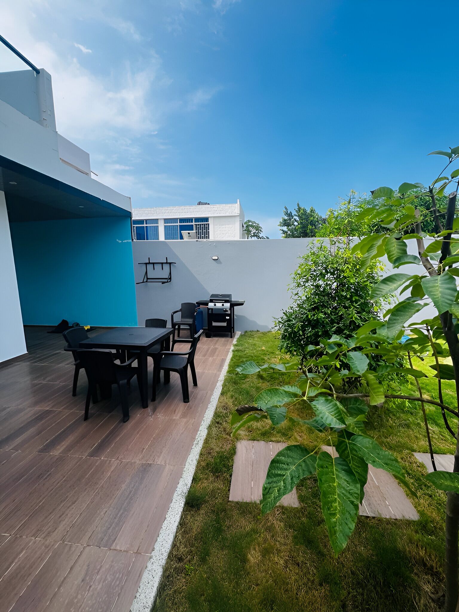 Un patio al aire libre con una mesa y sillas, un pequeño jardín con un sendero, y un edificio moderno al fondo.