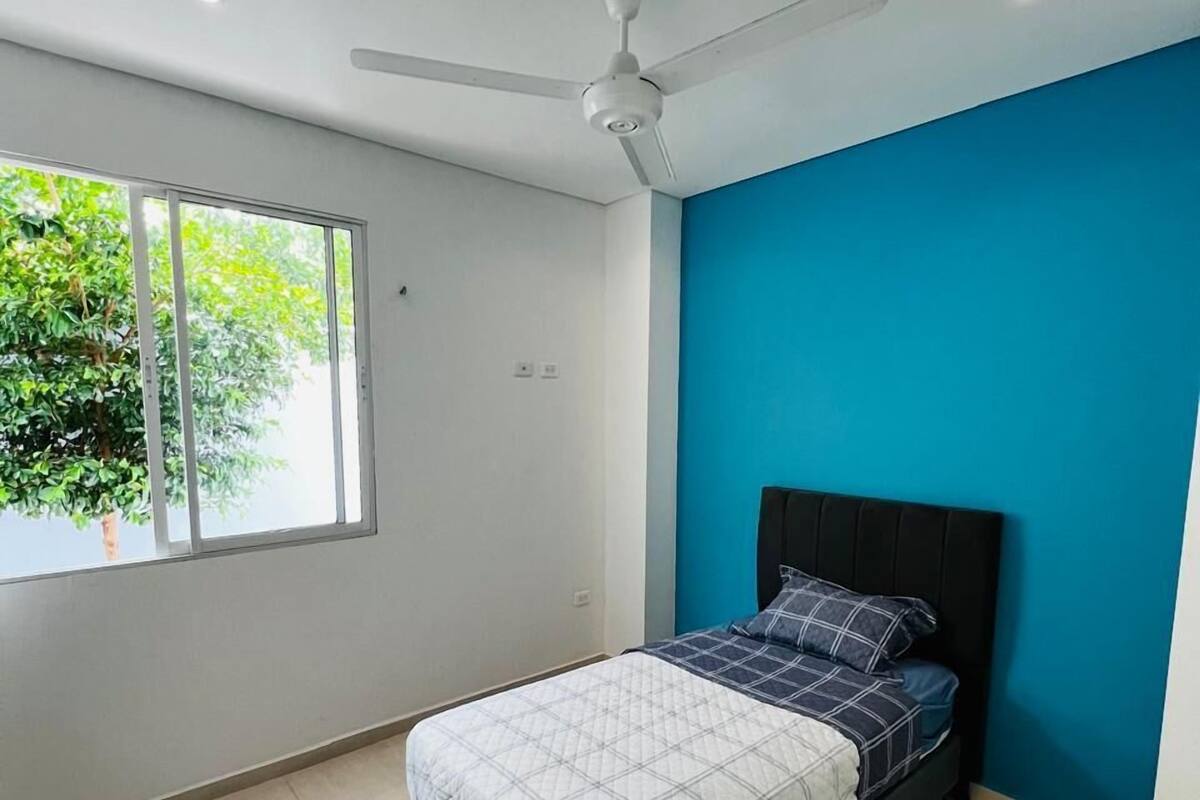 A bedroom with a bed, a ceiling fan, a window with a view of greenery, and a blue accent wall.