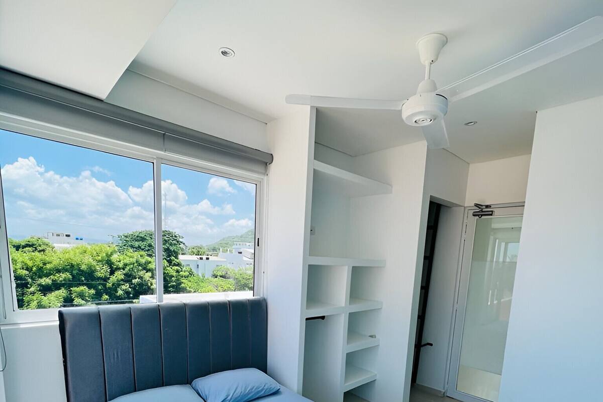 A bedroom with a bed, a ceiling fan, a window with a view of greenery and buildings, and a built-in shelving unit.