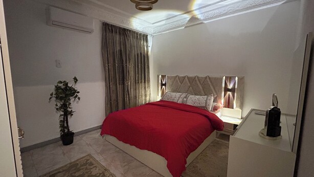 Un dormitorio con cama, mesita de noche, lámpara, planta en maceta y ventana con cortinas.