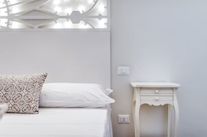 Una cama con ropa de cama blanca y un cojín estampado, una mesita de noche con cajón, e interruptor de luz fijado a la pared.