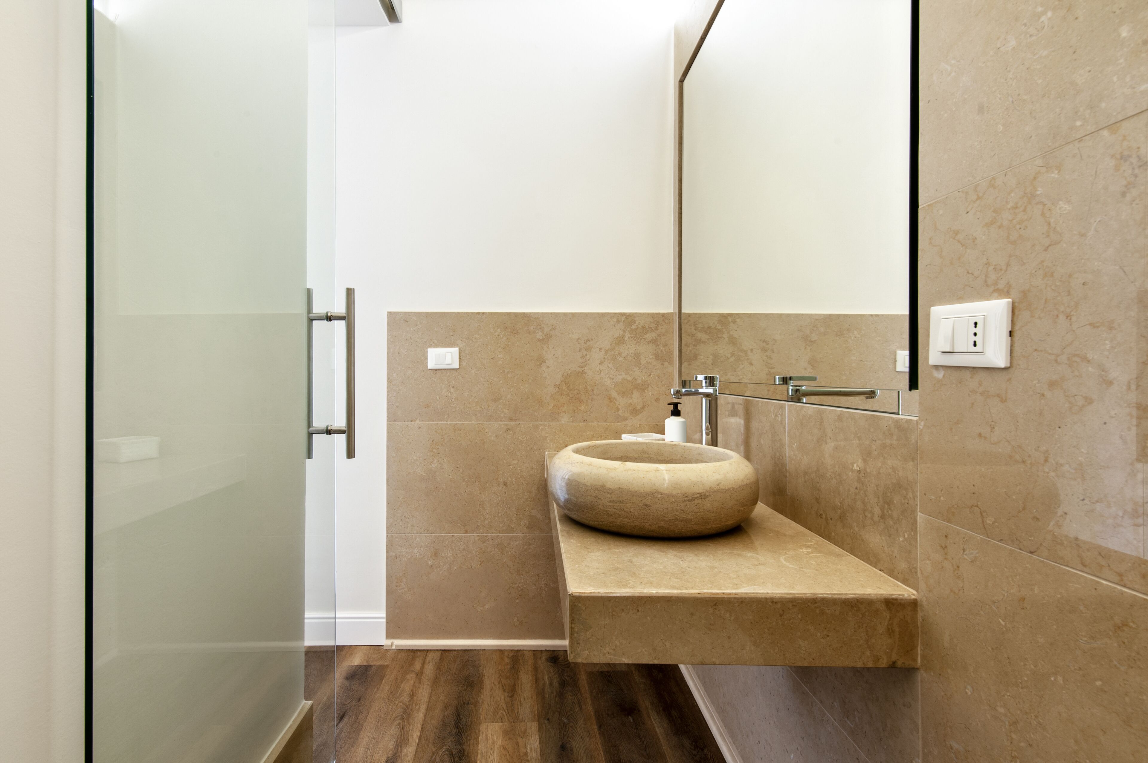 Un baño moderno con cabina de ducha de vidrio, lavamanos de piedra color beige y un espejo grande.