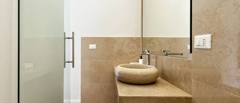 Un baño moderno con cabina de ducha de vidrio, lavamanos de piedra color beige y un espejo grande.
